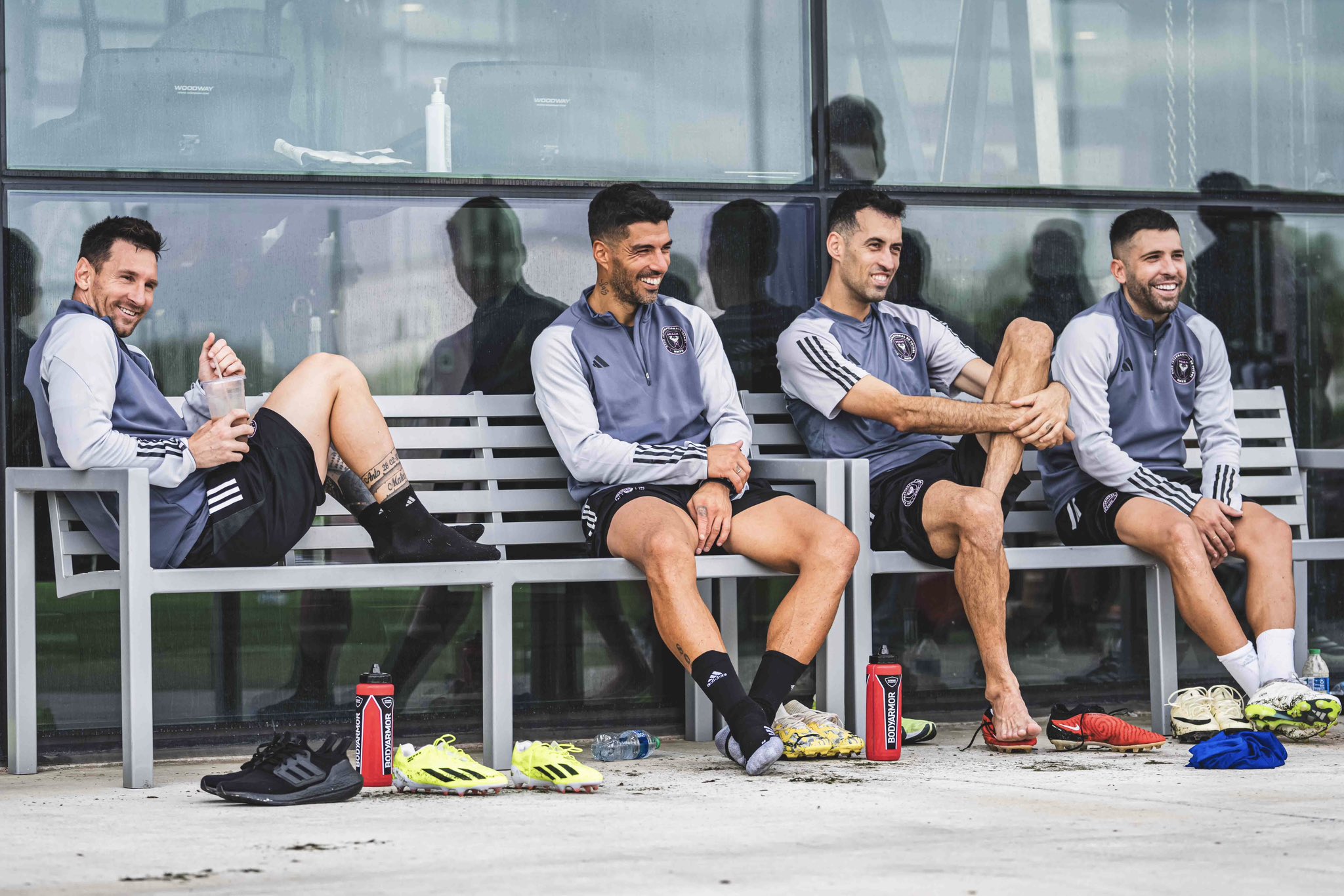 Lionel Messi, Luis Suarez, Sergio BUsquets, and Jordi Alba sitting on grey benches at training. 