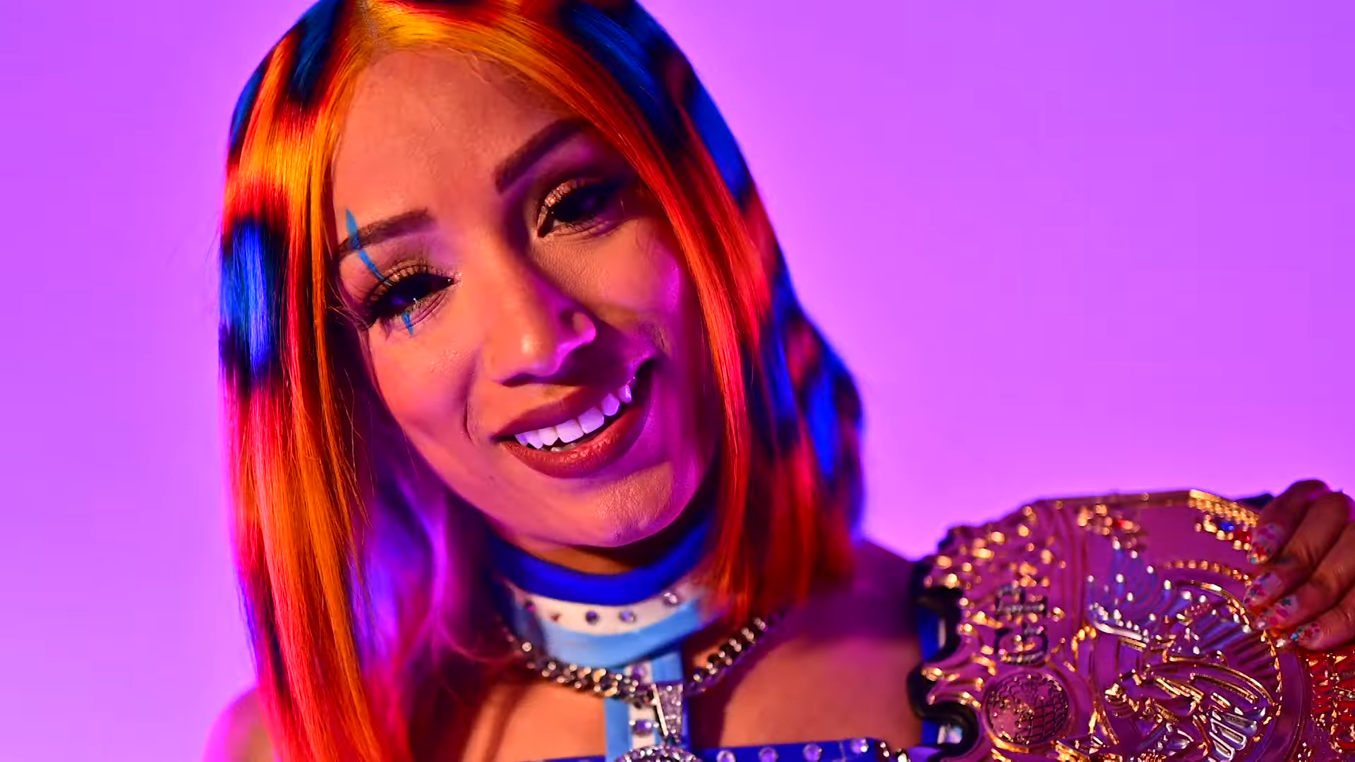 Mercedes Mone, with blue and orange hair, holding the gold IWGP Women's Championship in front of a purple wall.