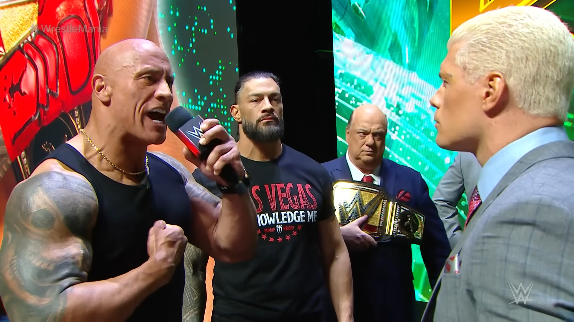 A screenshot from WWE's WrestleMania 40 Press Conference. The Rock is holding a microphone, talking to Cody Rhodes. Roman Reigns stands beside him, with Paul Heyman holding the Undisputed WWE Championship.